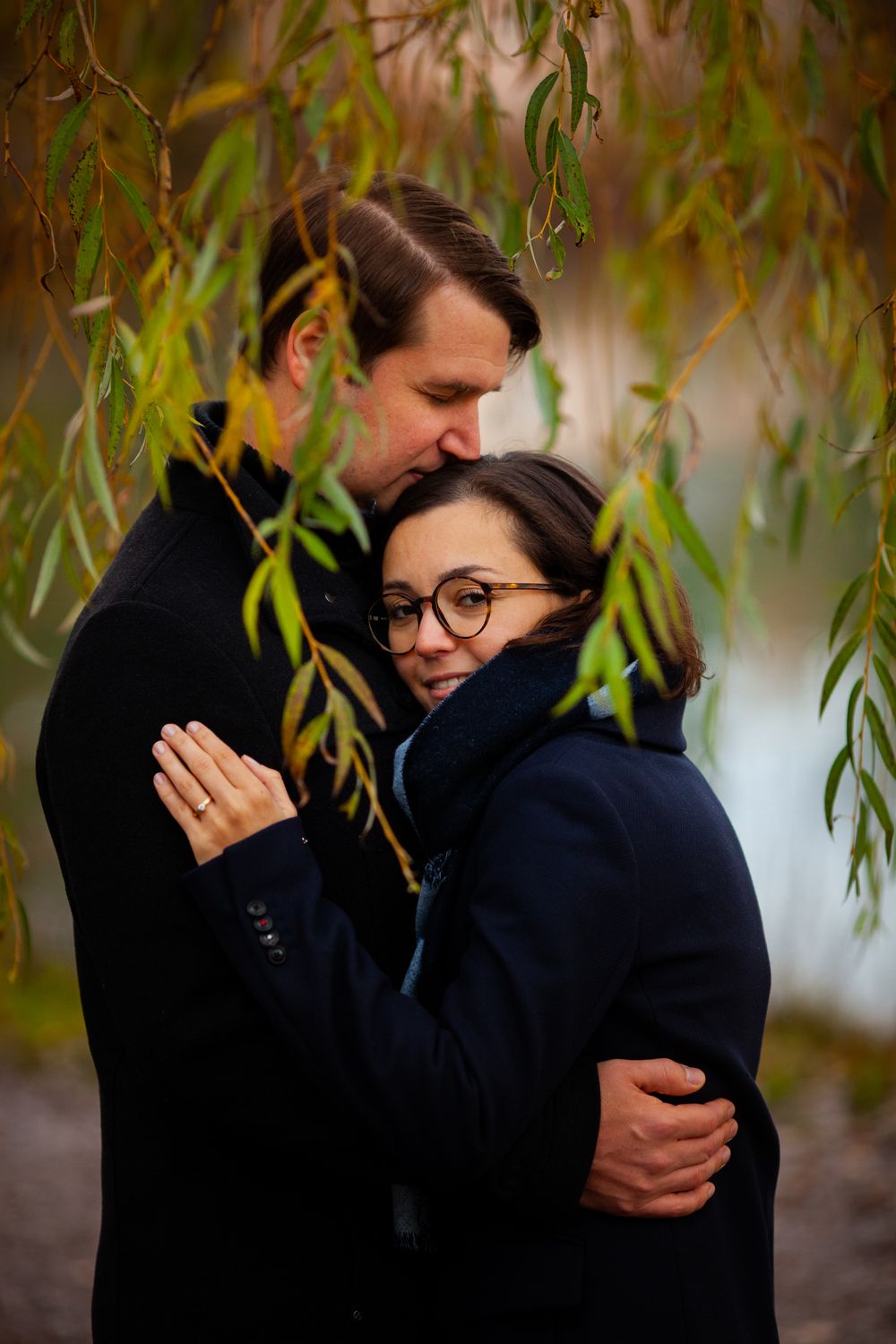 A couple embraces under hanging willow branches during an autumn engagement photo session.