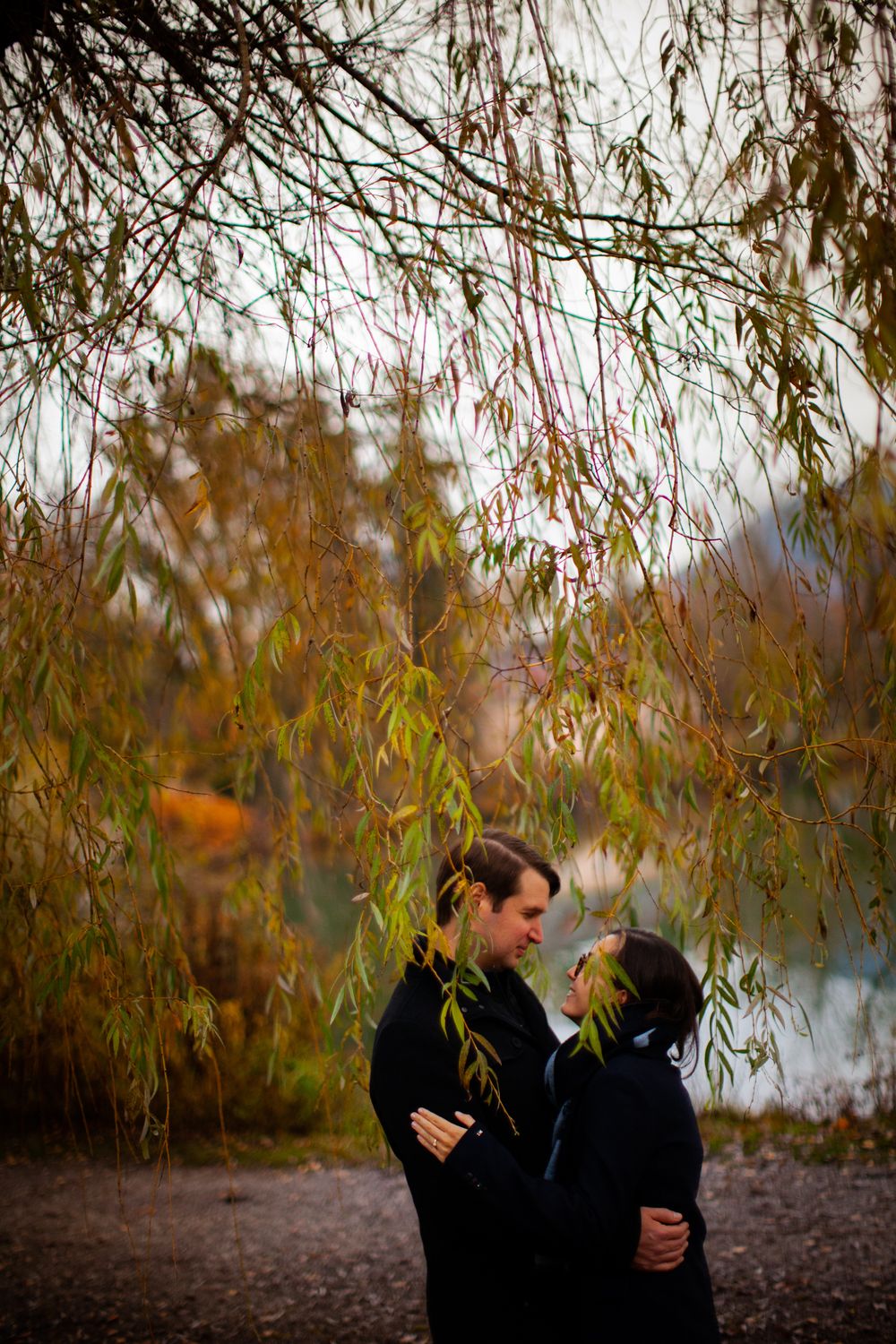 A romantic couple shares an intimate moment under weeping willow branches beside a lake at sunset.