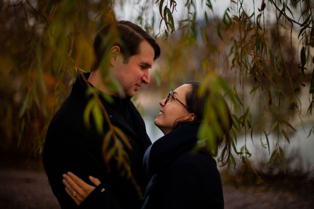 Romantic couple embraces under willow trees by a serene lake at sunset, creating an intimate outdoor moment.