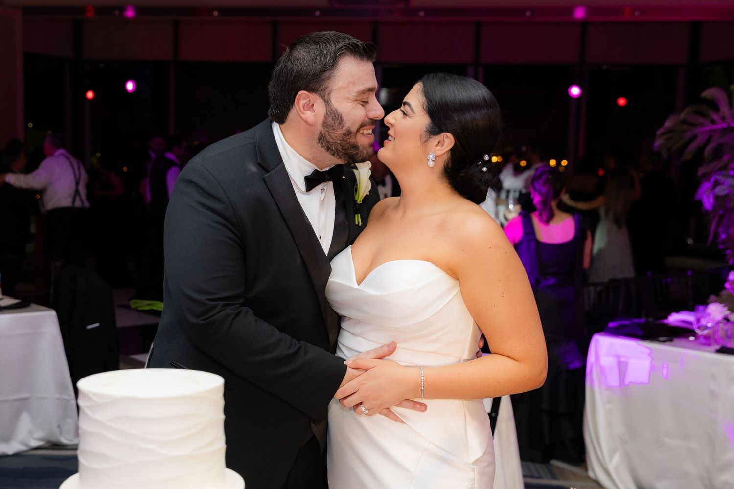 Newlyweds share intimate moment by wedding cake during reception with purple lighting.