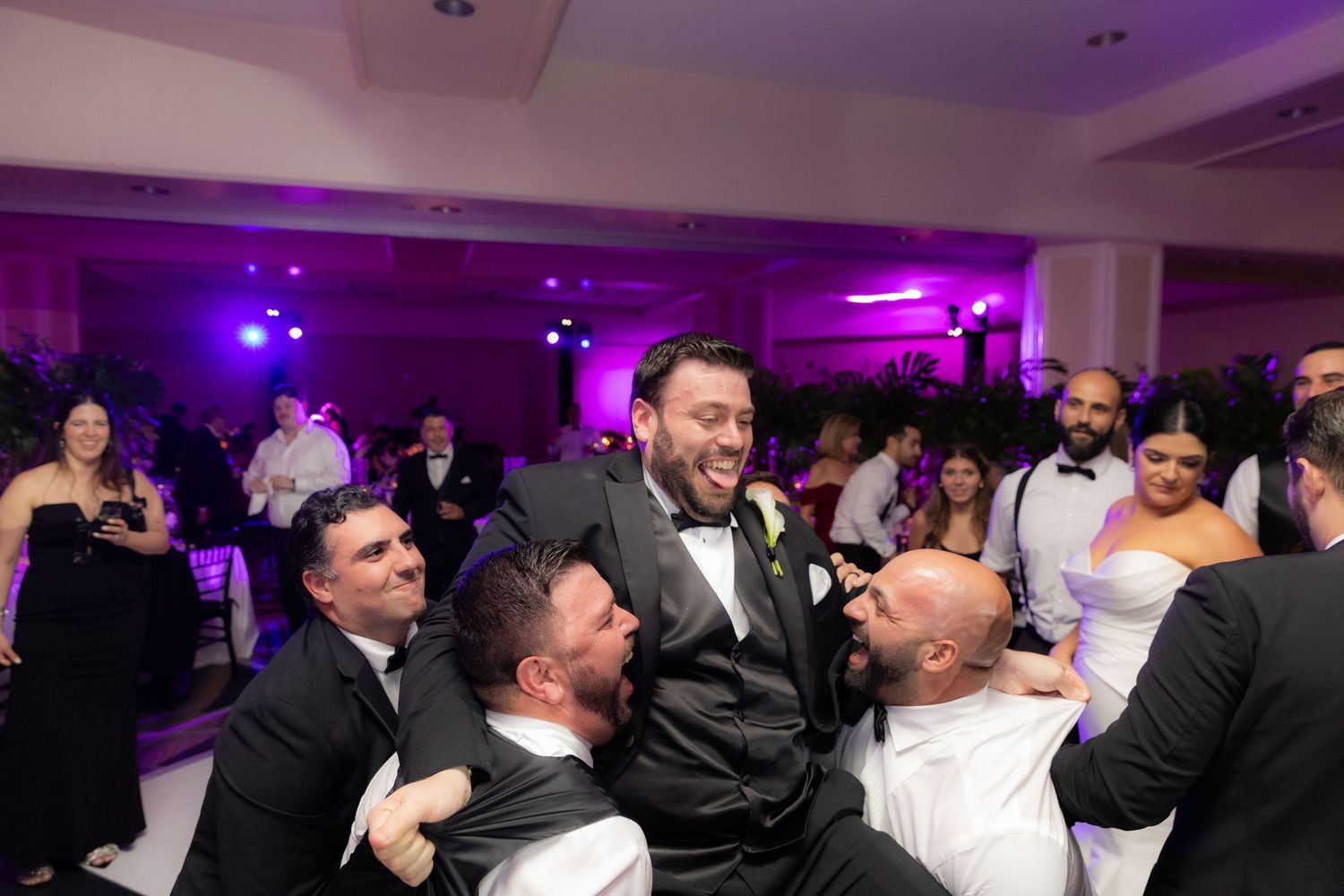 A group of party guests dancing together on a dance floor illuminated by purple lighting at an evening celebration.