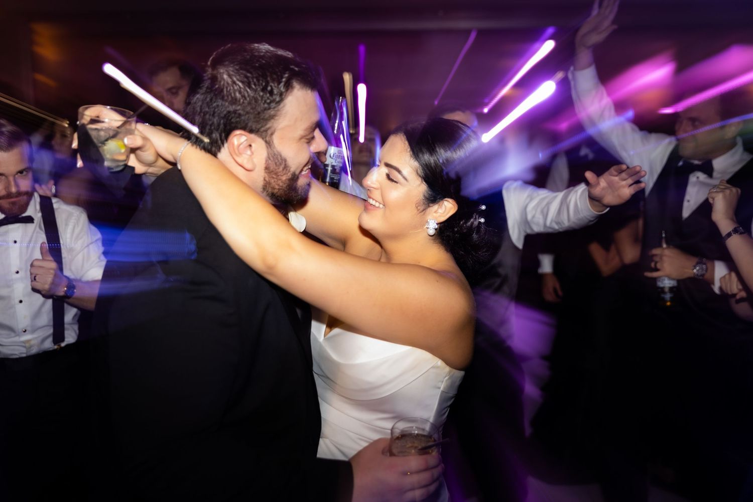 Wedding guests celebrate and dance together on a lively dance floor illuminated by purple lighting.