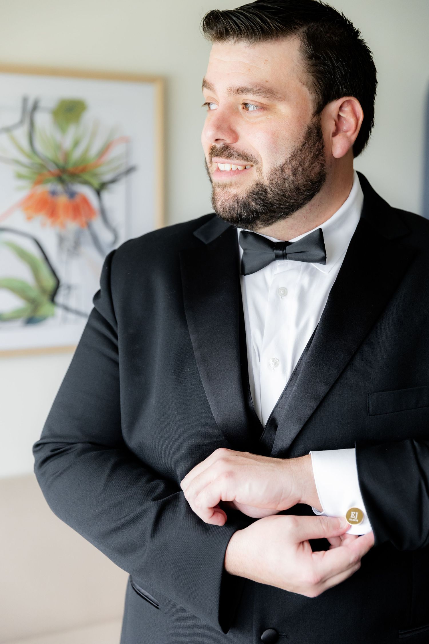 A groom in a classic black tuxedo and bowtie smiles during wedding preparations.