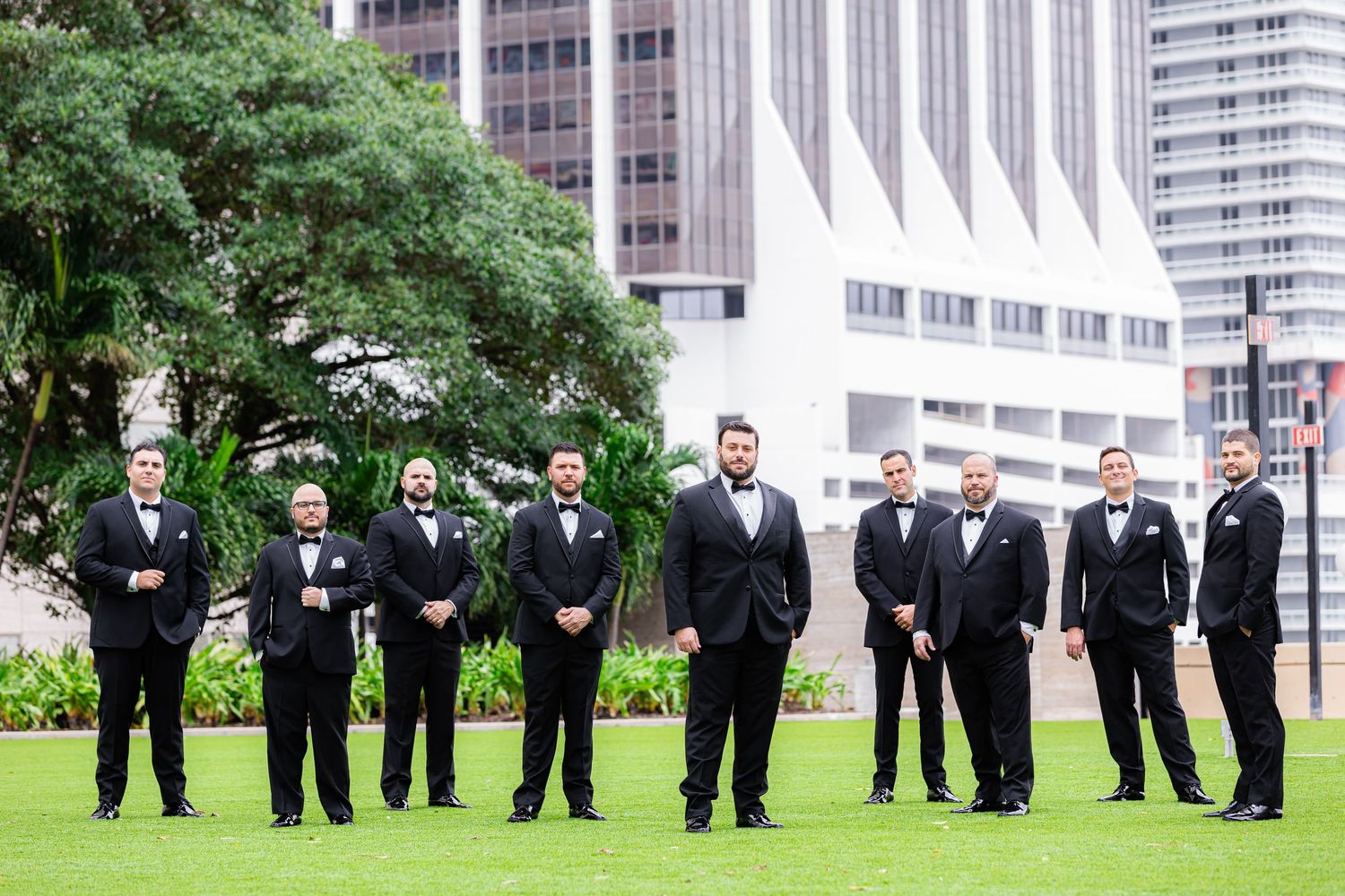 Groomsmen in black tuxedos stand in a row on green lawn with modern buildings in background.