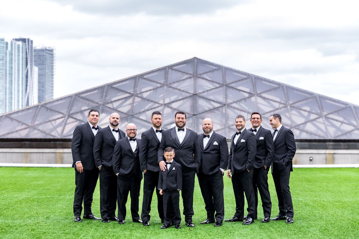 Groomsmen in black suits and ties pose together outdoors in front of a modern building.