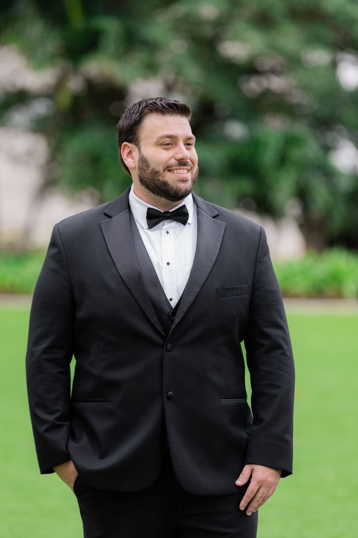 Well-dressed person in a black tuxedo and bow tie standing on grass outdoors.