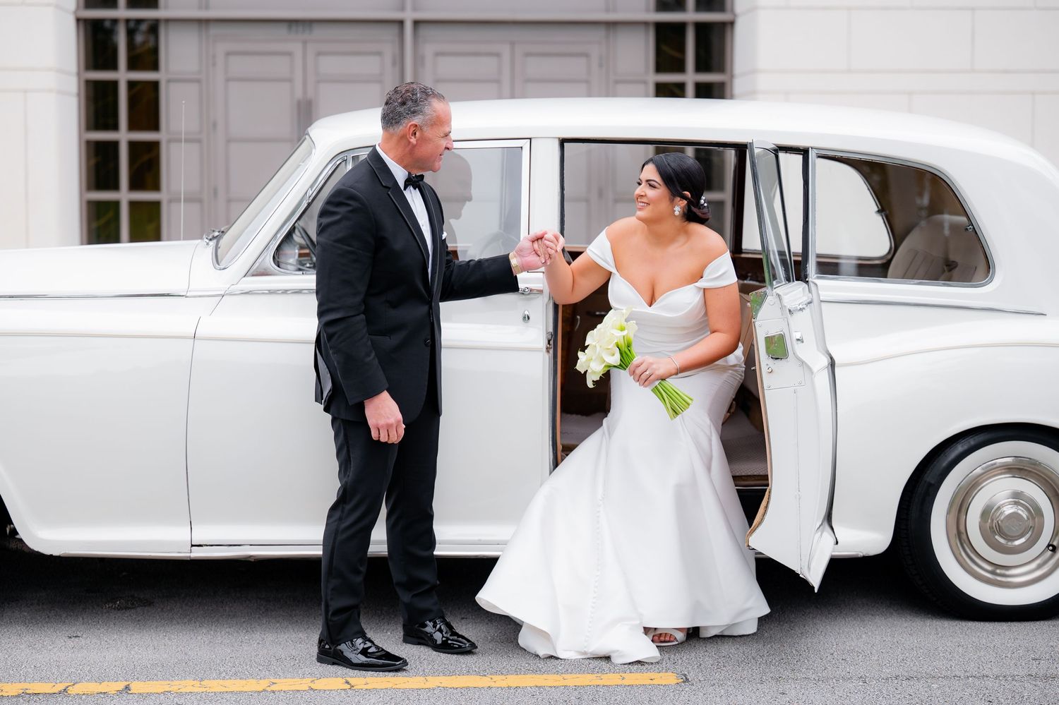 A classic white vintage car serves as elegant wedding day transportation outside a stately venue.
