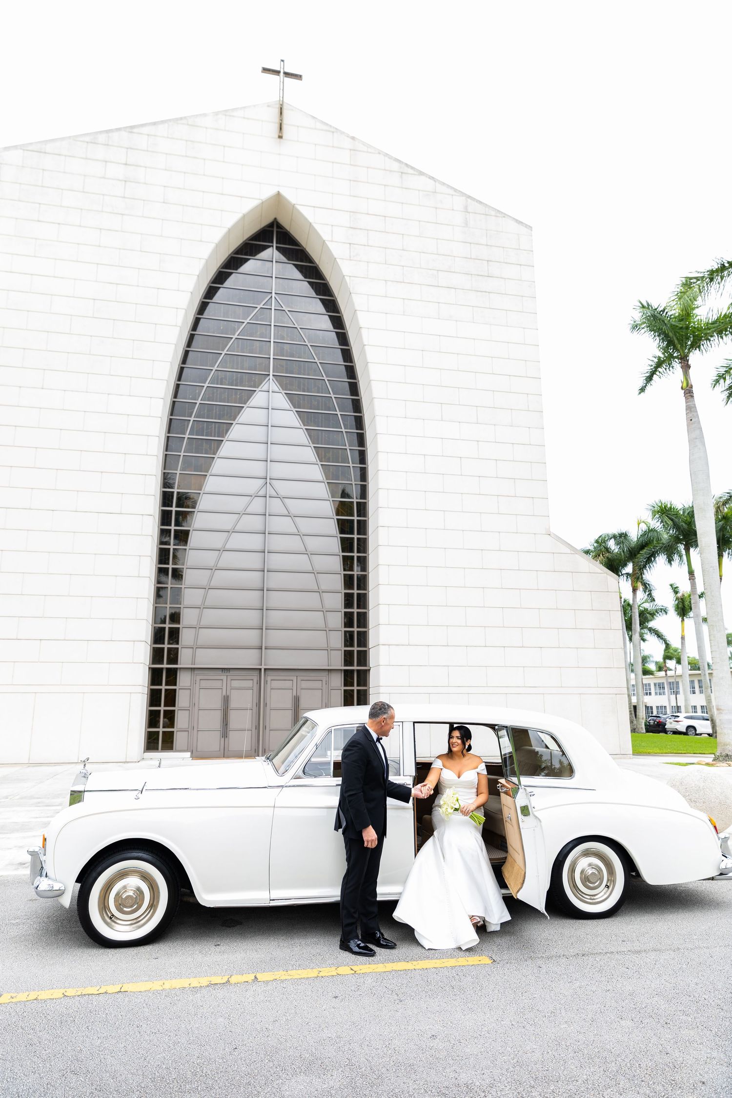 Classic white vintage car parked in front of a modern church with palm trees.