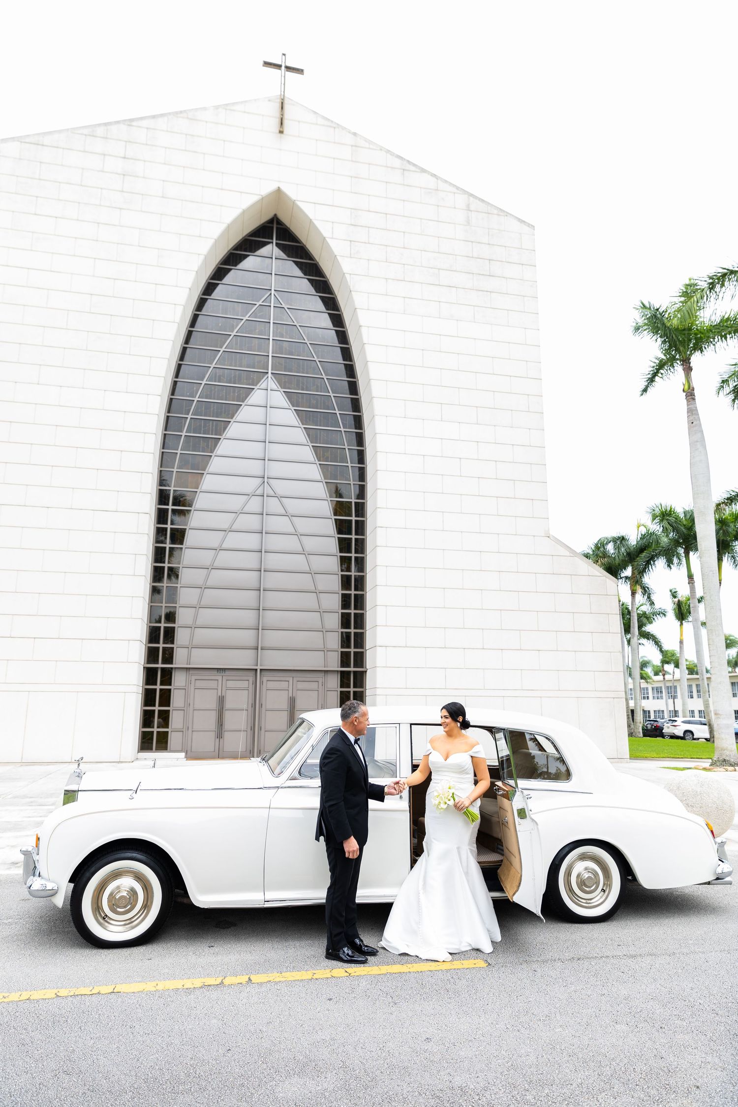 White vintage Rolls Royce parked outside a church with Gothic arched windows.