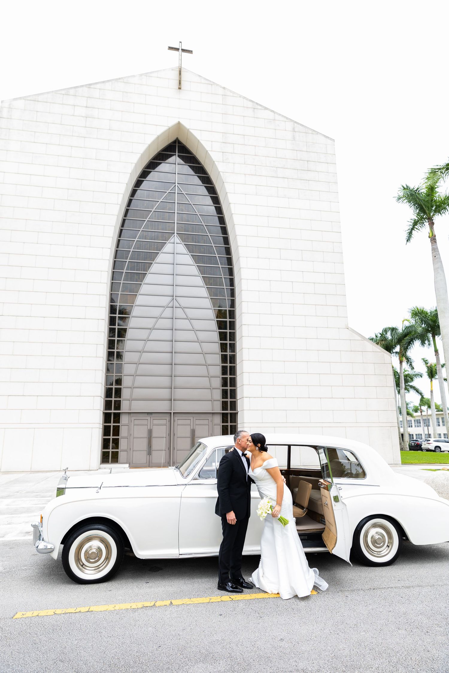 A wedding couple poses outside a white church with a vintage white Thunderbird car parked nearby.