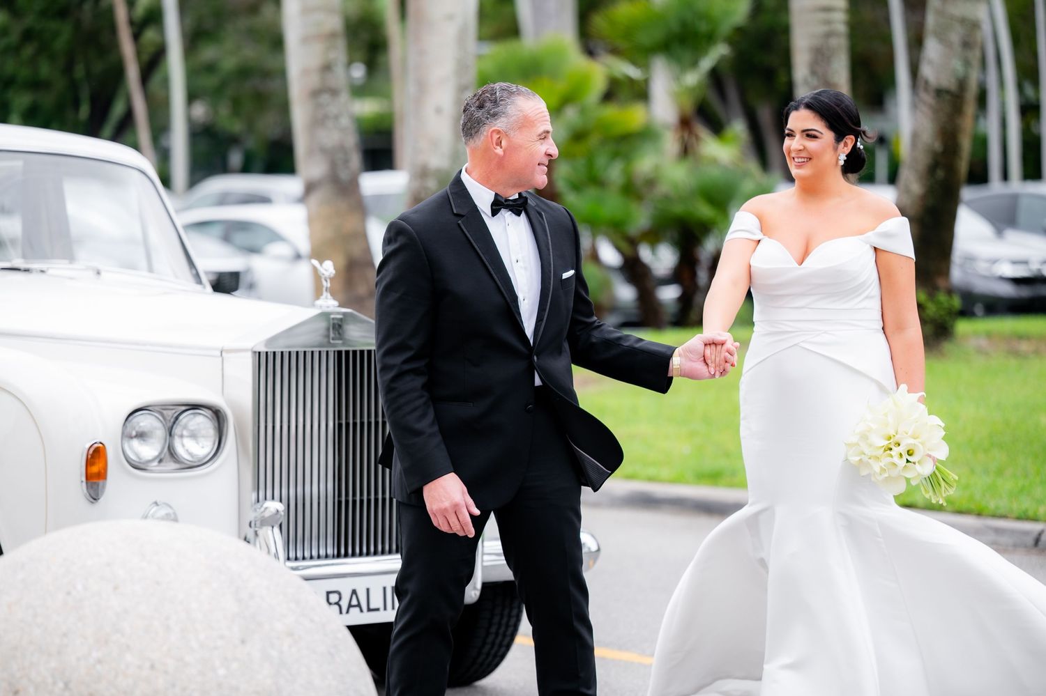 Elegant bridal couple walking beside classic Rolls Royce with palm trees in background.