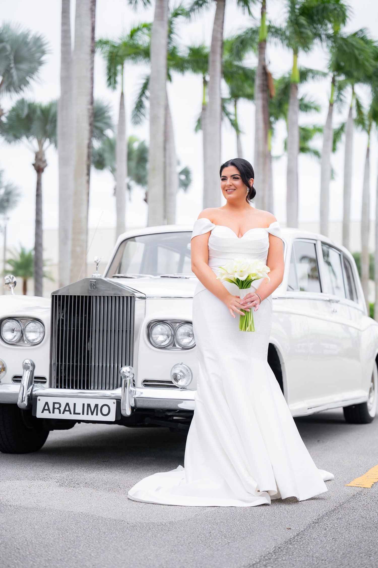 White Rolls-Royce wedding cars line a palm tree-lined driveway with bride in foreground.