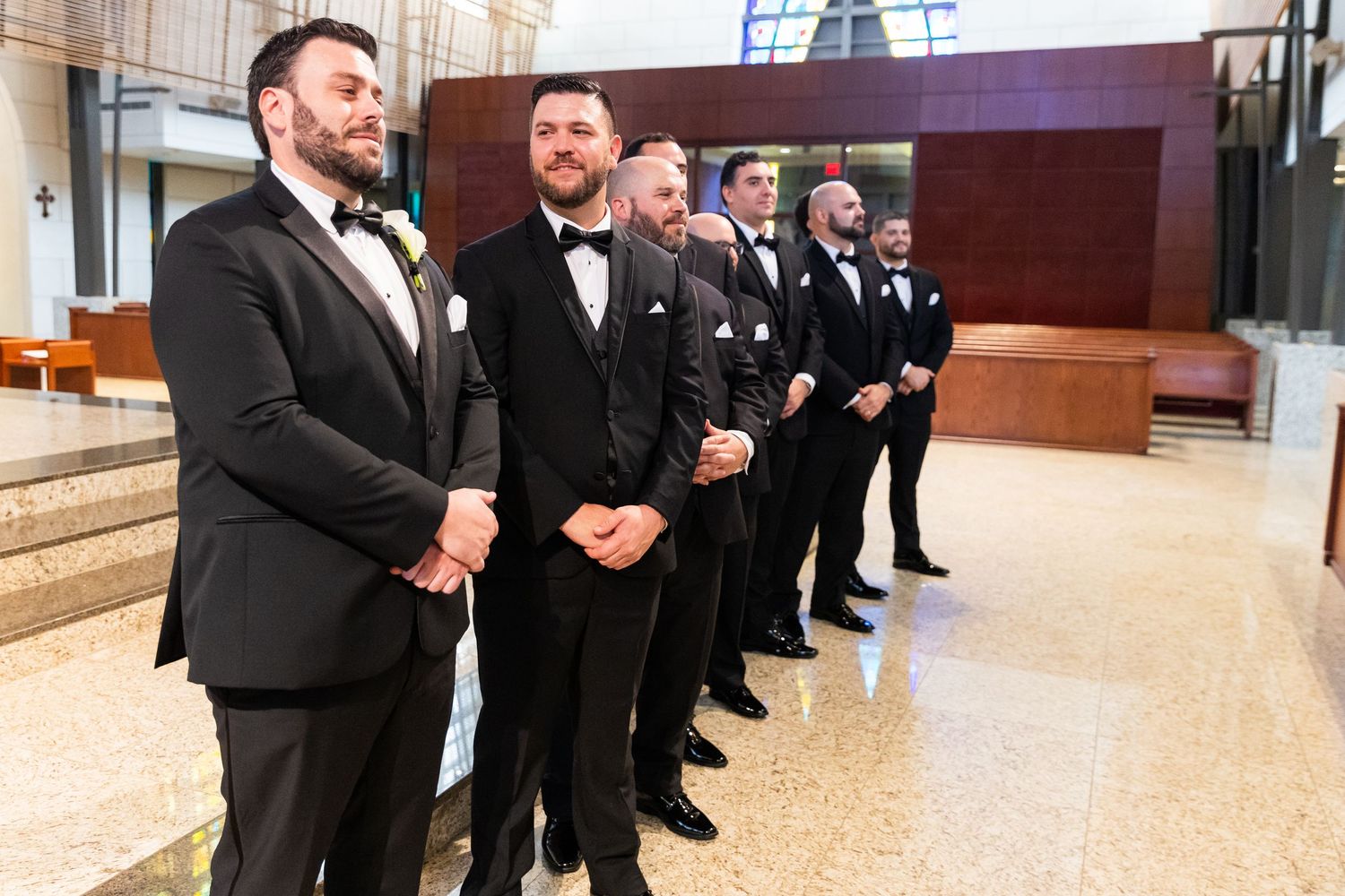 Wedding party members in black tuxedos line up in an indoor venue.