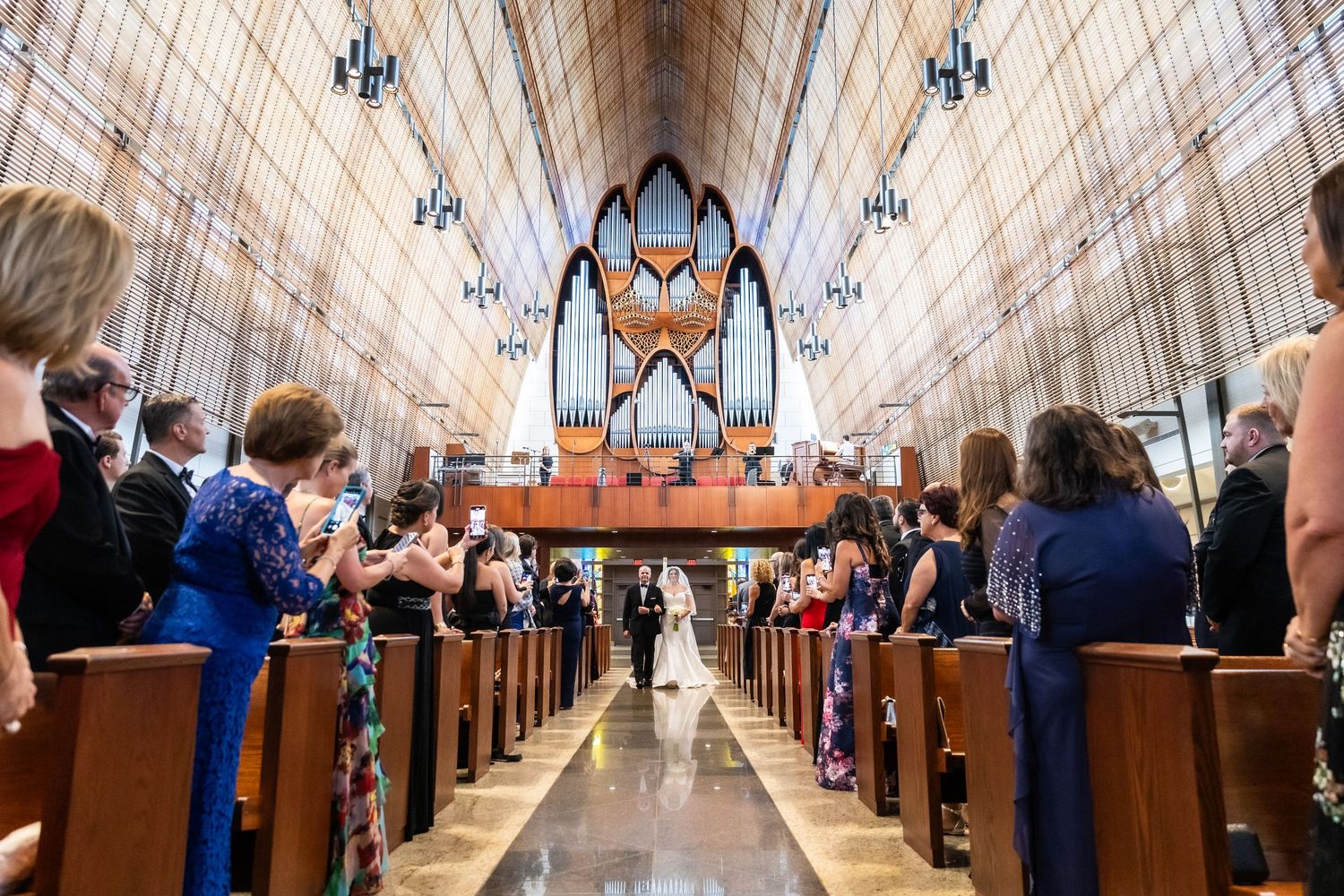 Wooden pews line the aisle of a grand cathedral with soaring Gothic arches and stained glass windows.
