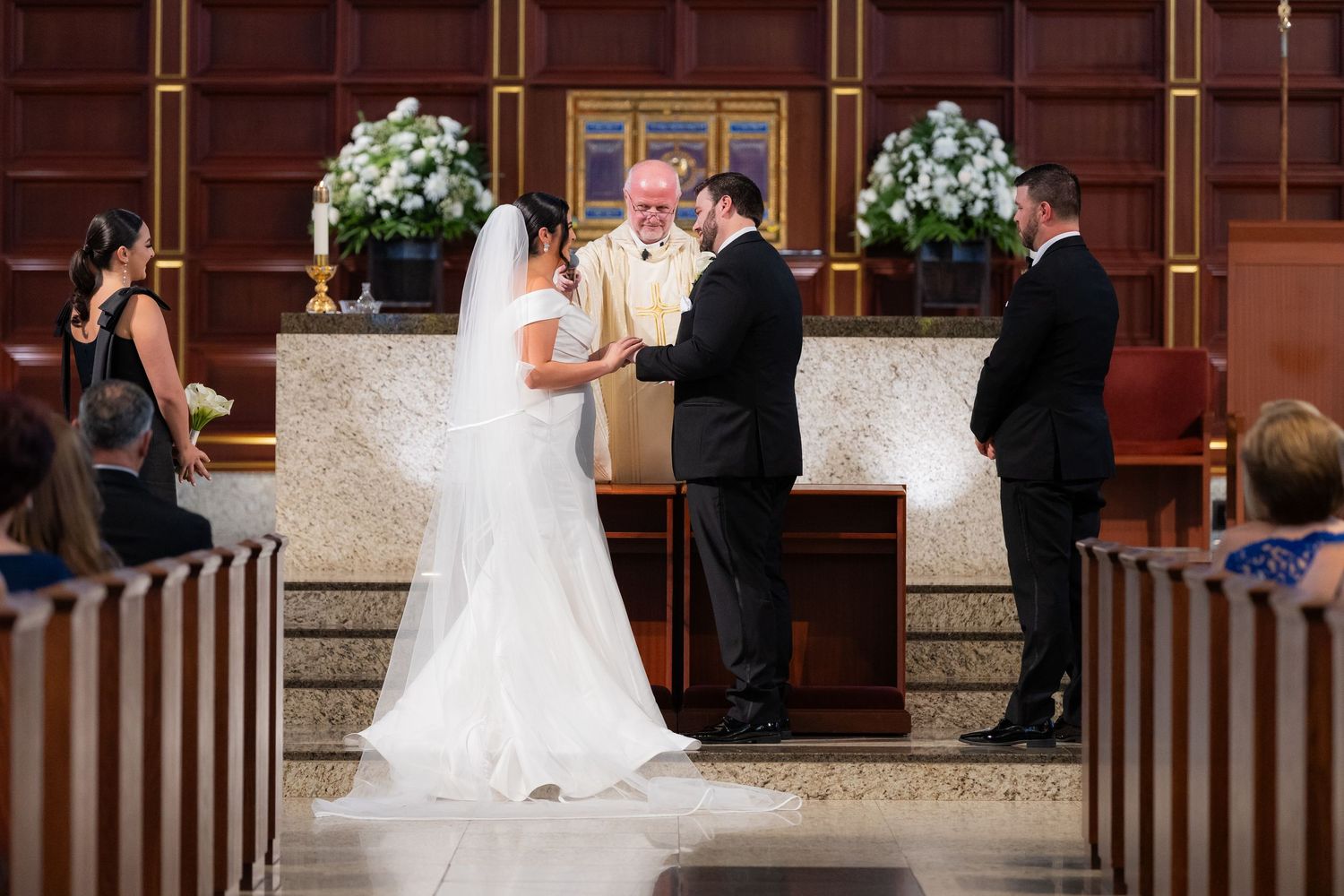 Wedding ceremony in progress at church altar with white floral arrangements.