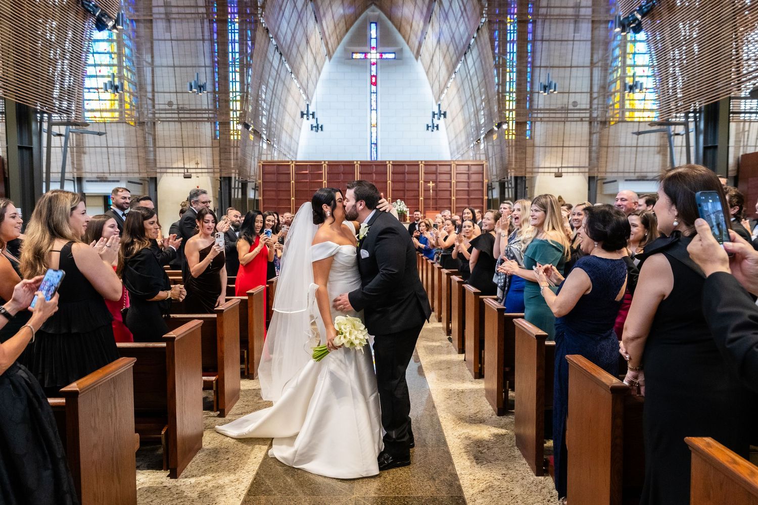 Wedding guests stand in church pews as couple processes down aisle decorated with flowers.