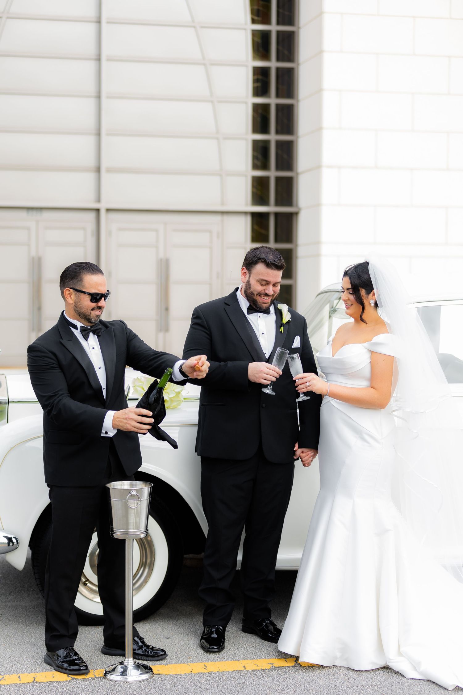 Wedding party members in formal attire peek around pillars in a playful pose sequence along a white building exterior.