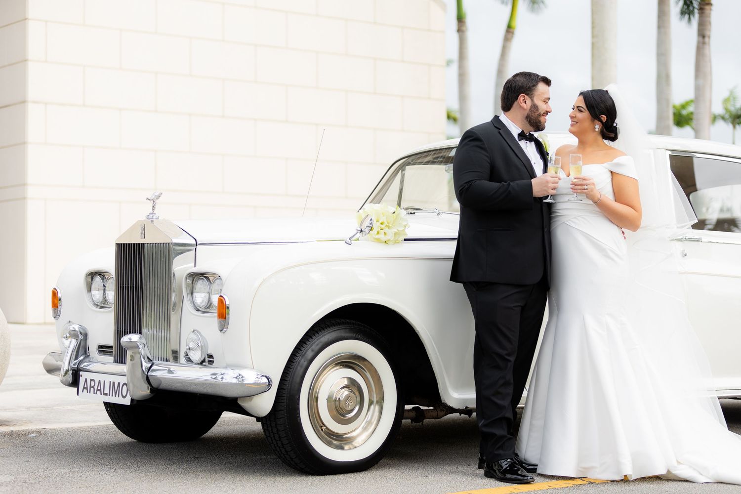 Beautiful wedding portraits with vintage white car and elegant couple against white building.
