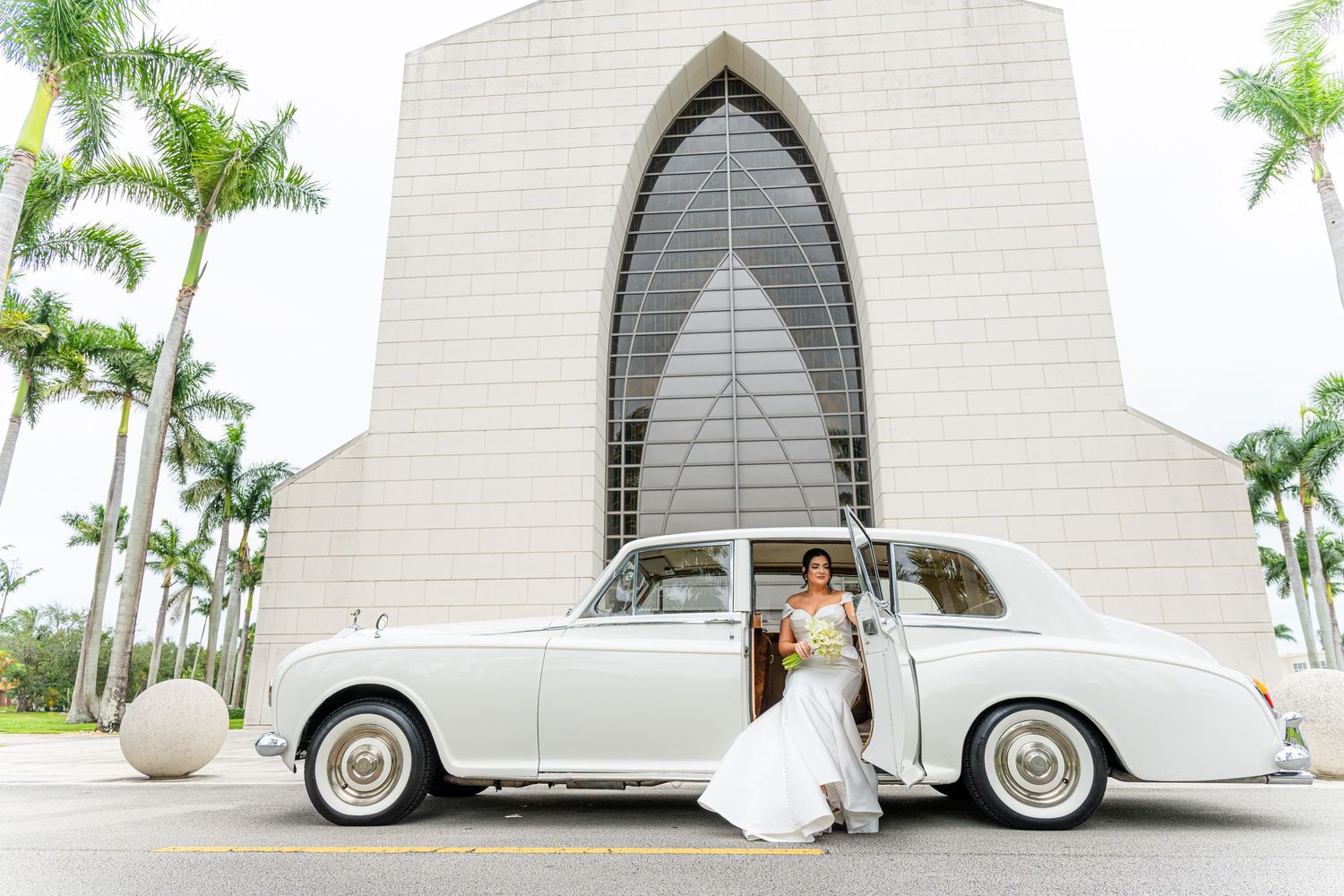 Modern white church exterior with vintage wedding car and palm trees.