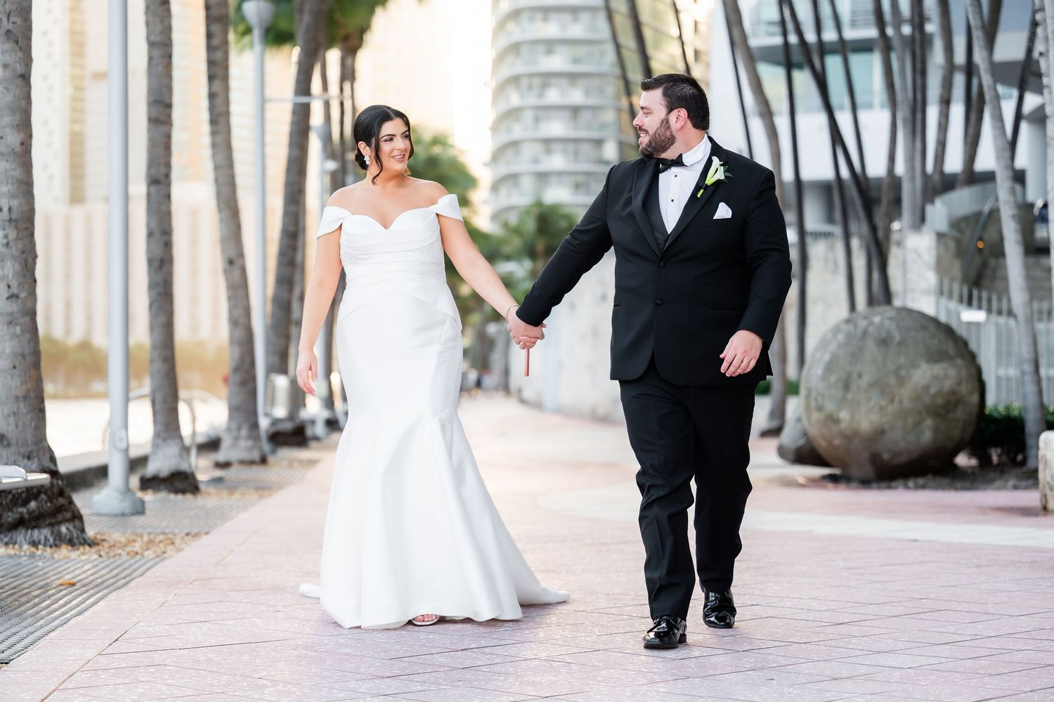 A couple walks along a palm-tree lined pathway in formal wedding attire.