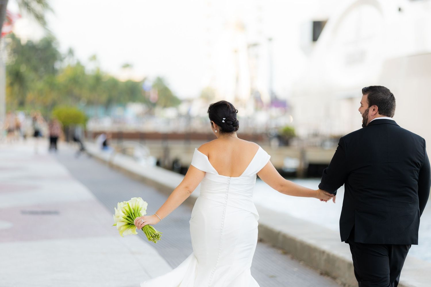 Wedding couple walking hand-in-hand on an outdoor path surrounded by palm trees.