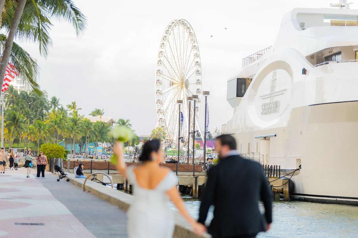 Romantic wedding couple walks along a beachfront path with palm trees and Ferris wheel in background.