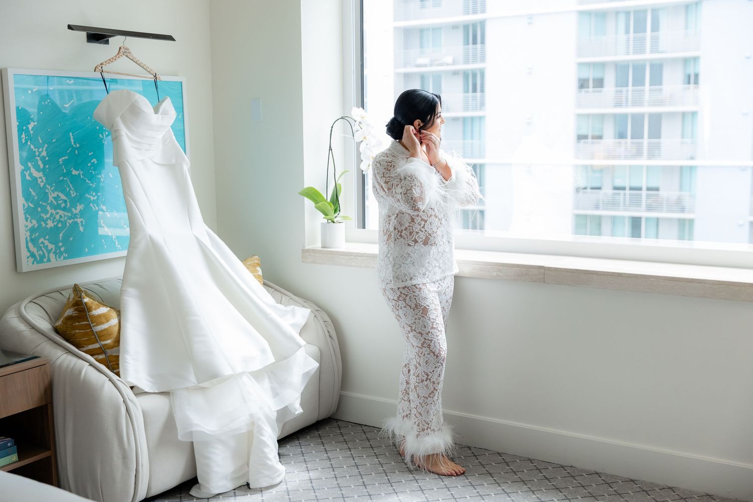 A bride in a white lace wedding dress poses by bright windows in an indoor setting.