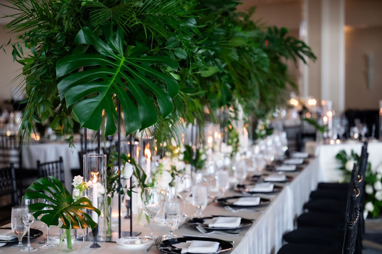 Elegant wedding reception table setting with lush greenery decorations and crystal glassware in a bright venue.