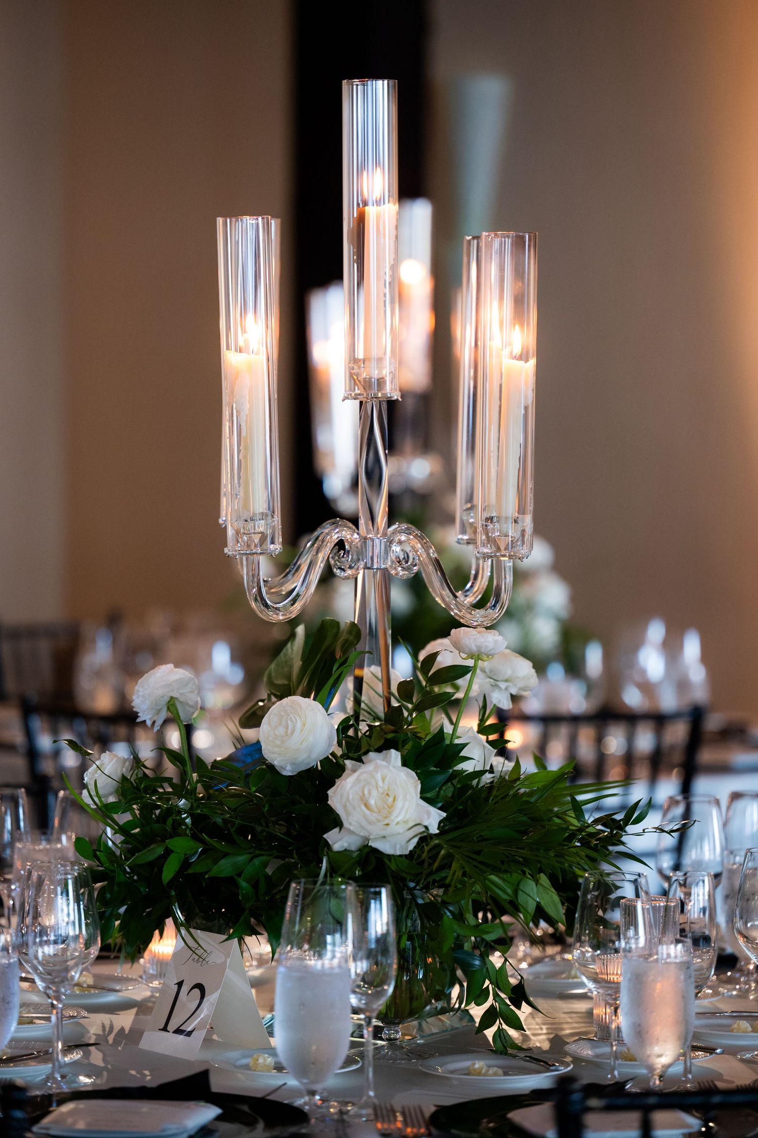 Elegant wedding reception tables with white floral centerpieces, crystal candelabras, and formal place settings in a banquet hall.