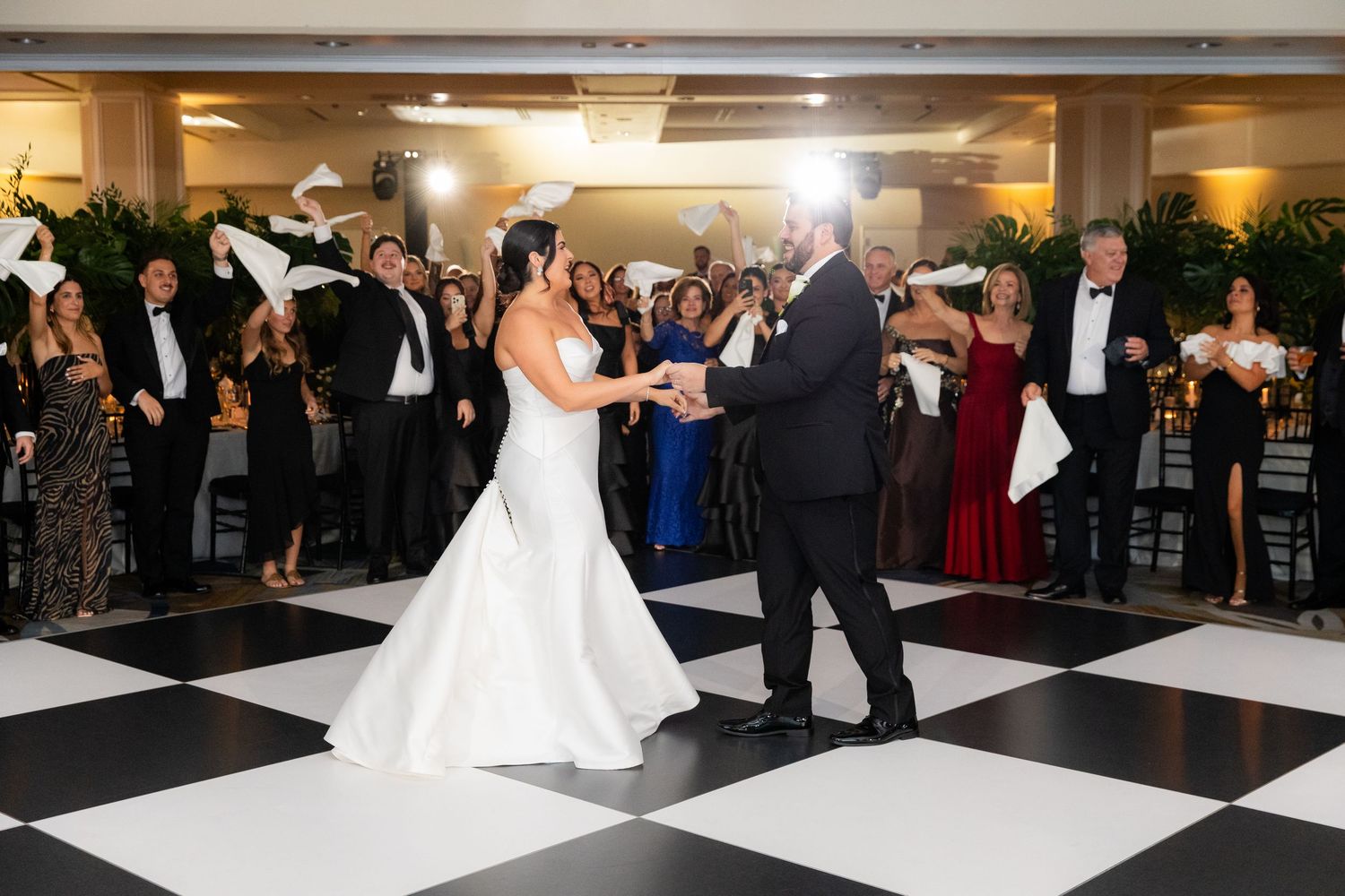 Newlyweds share their first dance surrounded by guests on a black and white checkered dance floor at an elegant reception.