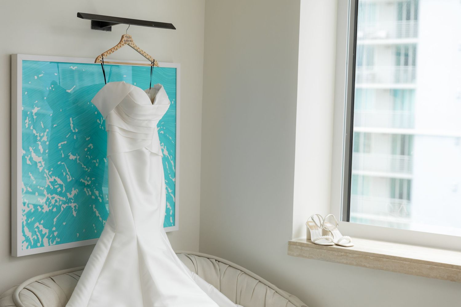 White strapless wedding gown hanging in room with turquoise wall art creating elegant contrast.
