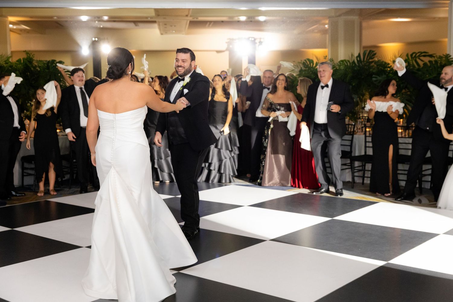 First dance of newlyweds on black and white checkered dance floor surrounded by guests.