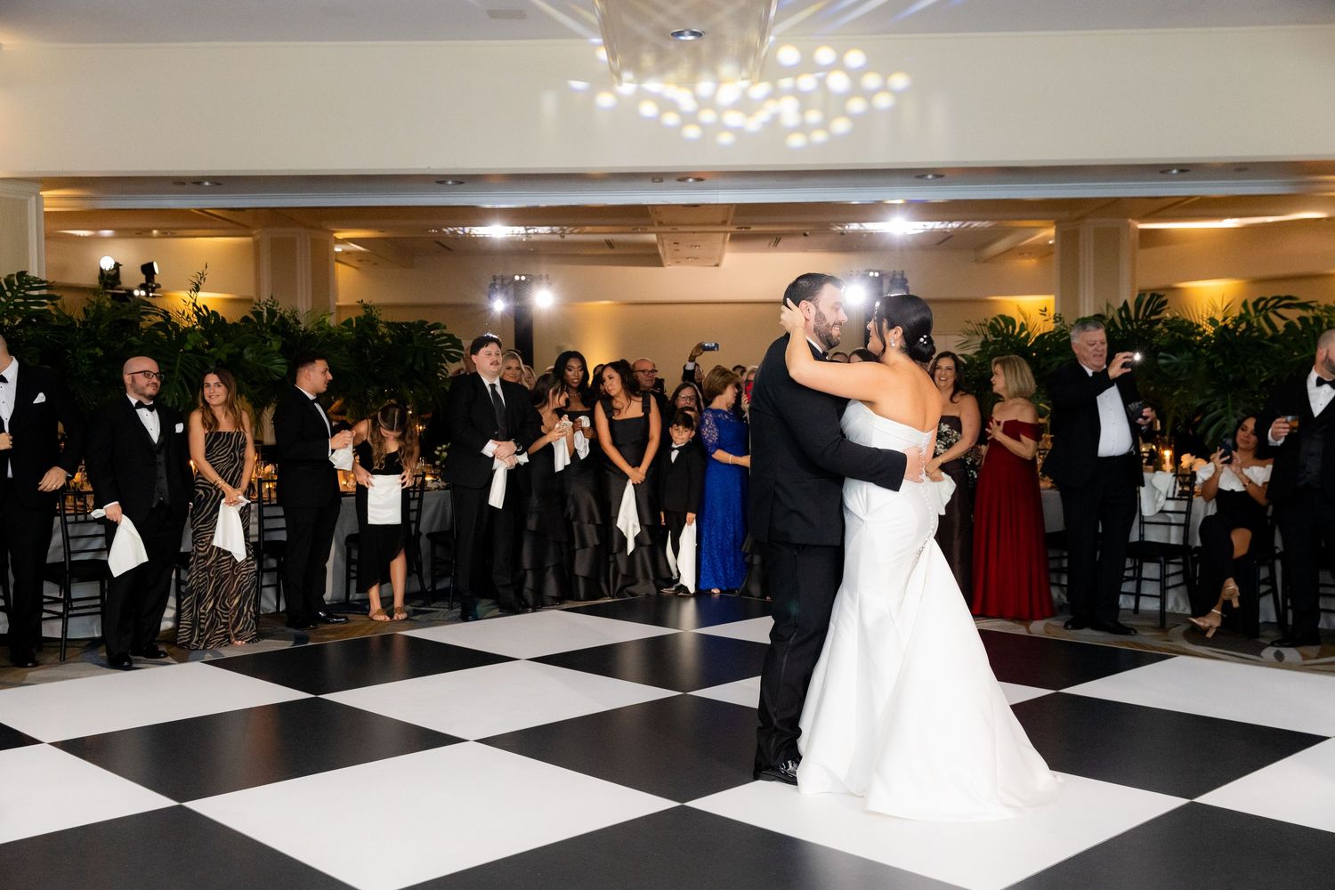 A first dance on a black and white checkered dance floor surrounded by wedding guests.