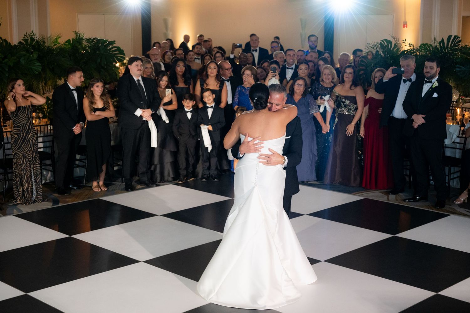 Newlyweds dance on a black and white checkered ballroom floor surrounded by wedding guests.