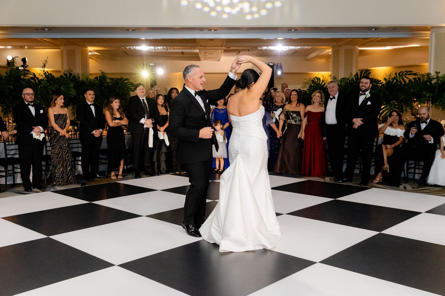 Couple performs their first dance on black and white checkered ballroom floor surrounded by guests.