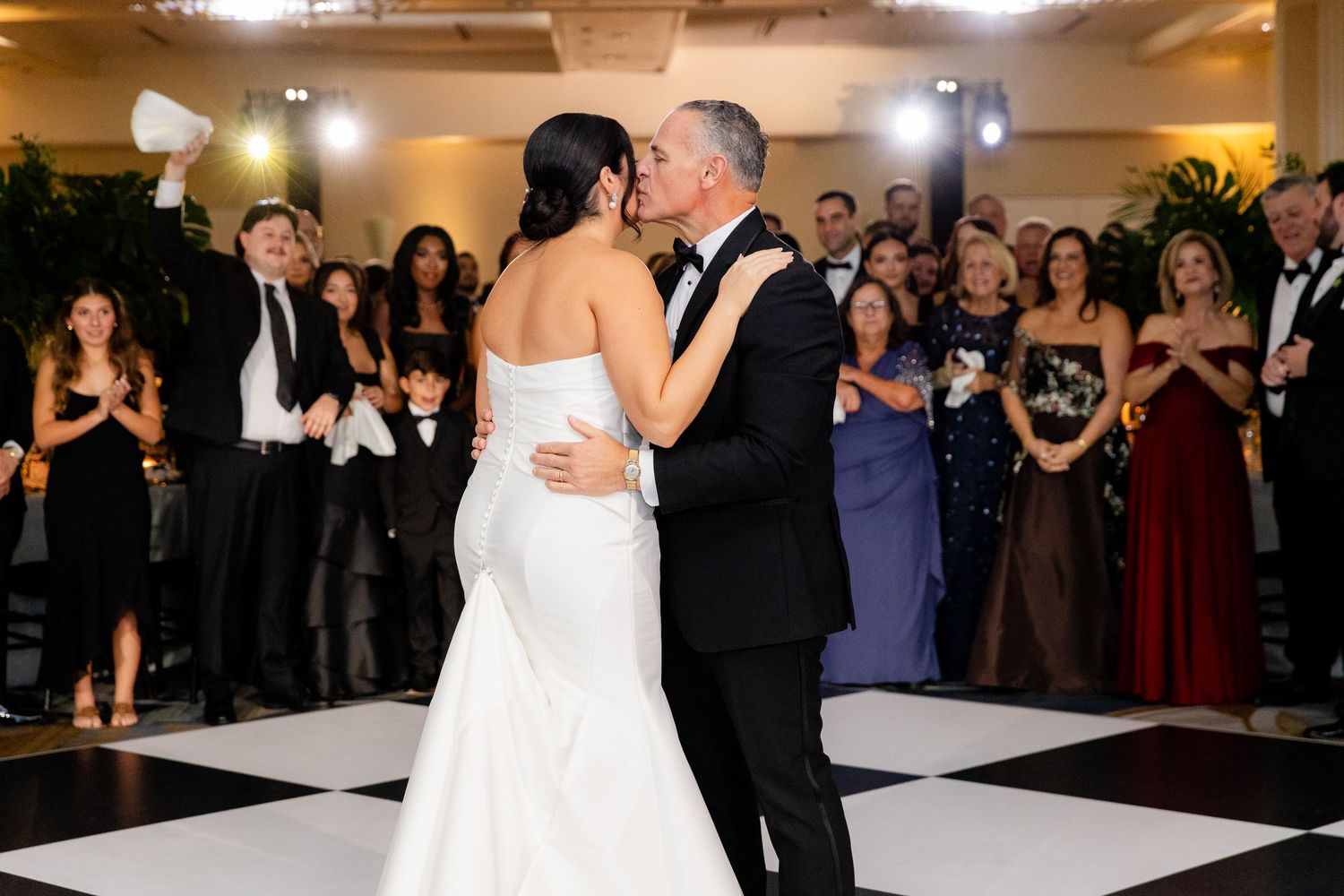 Intimate first dance kiss on black and white dance floor with guests looking on.