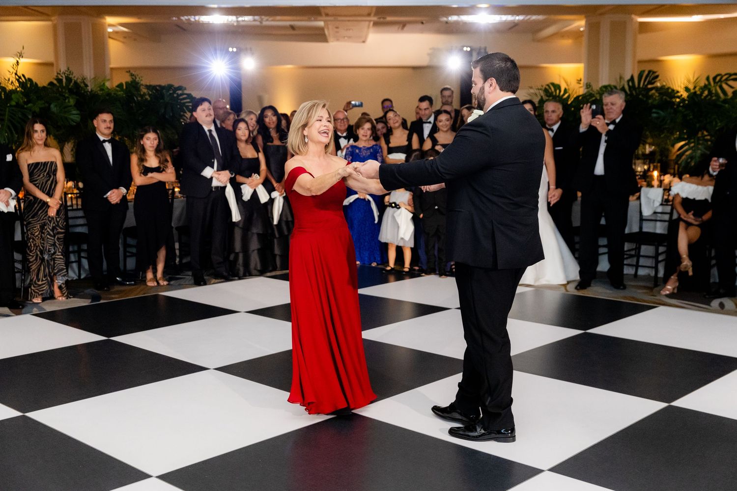 Wedding guests watching a couple dance on a black and white checkered floor with warm lighting.