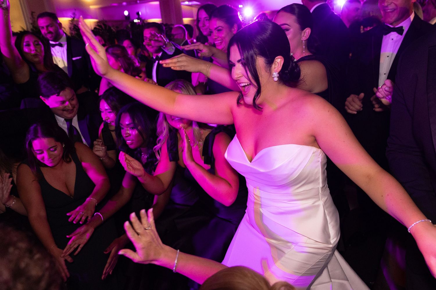 A lively crowd dances under vibrant purple lighting at a wedding reception or nightclub event.
