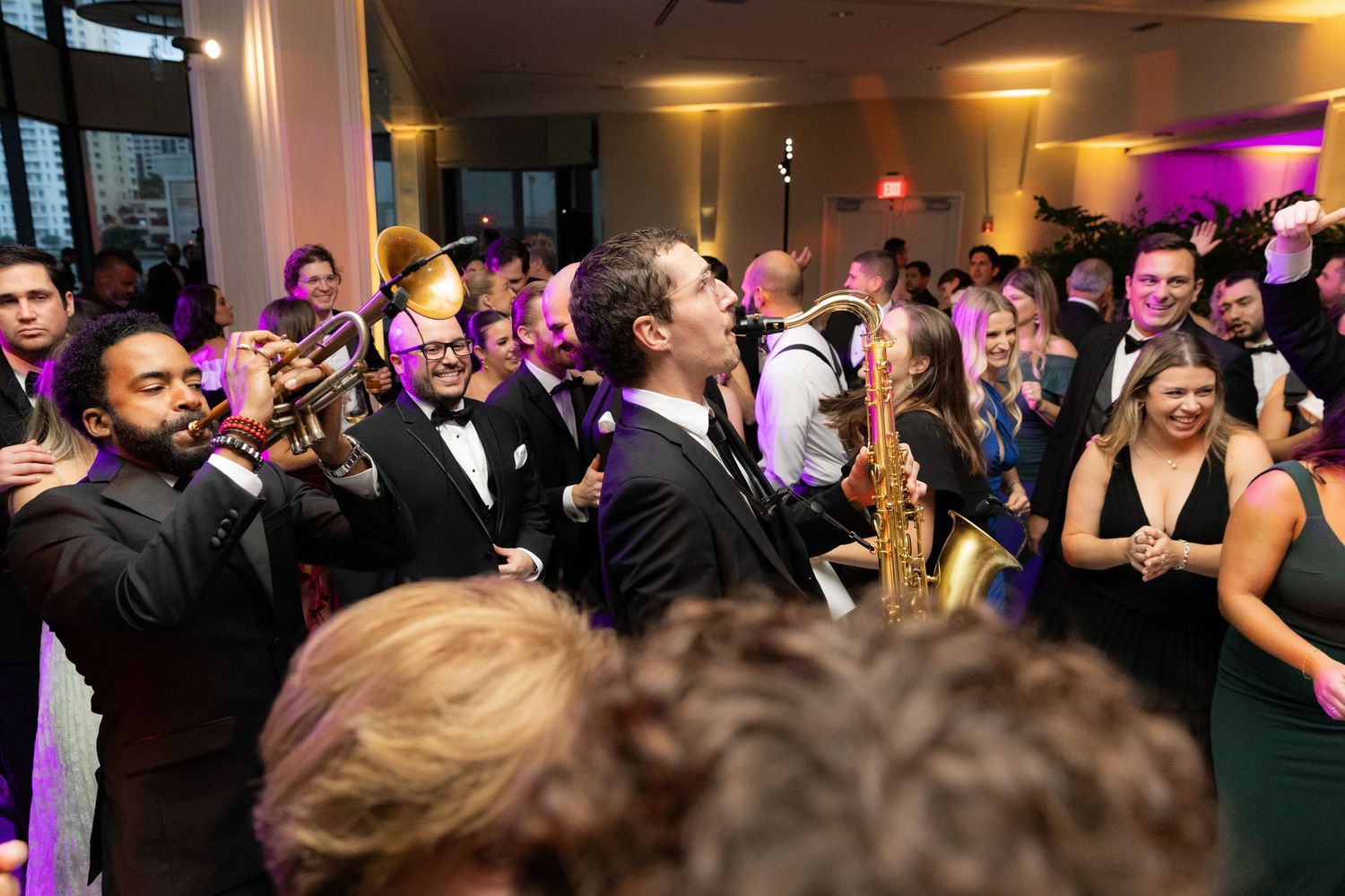 Wedding guests dance and celebrate on a dimly lit dance floor at an indoor reception venue.