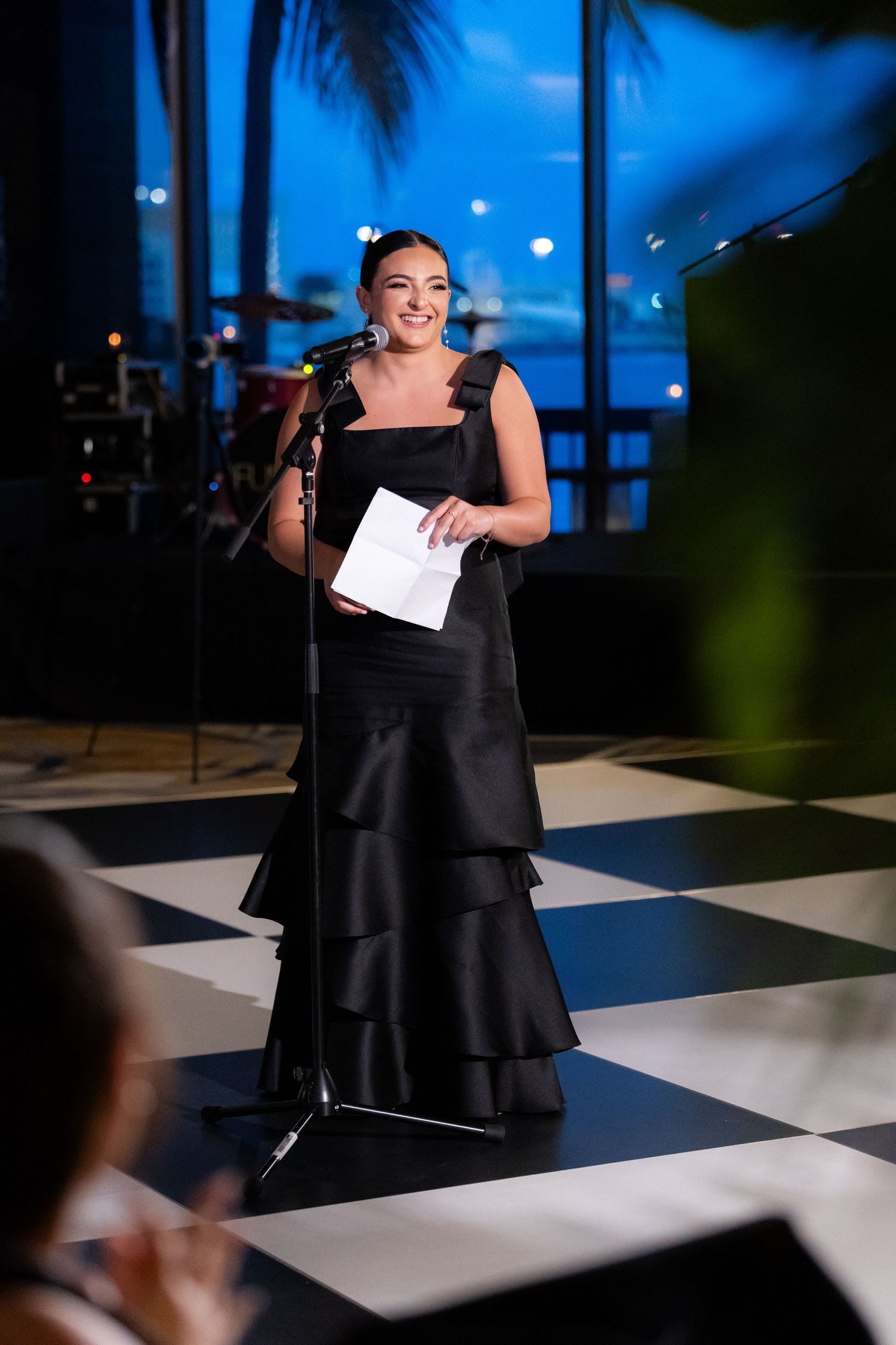 Speaker in elegant black tiered evening gown addresses audience from podium on checkered floor at evening event.