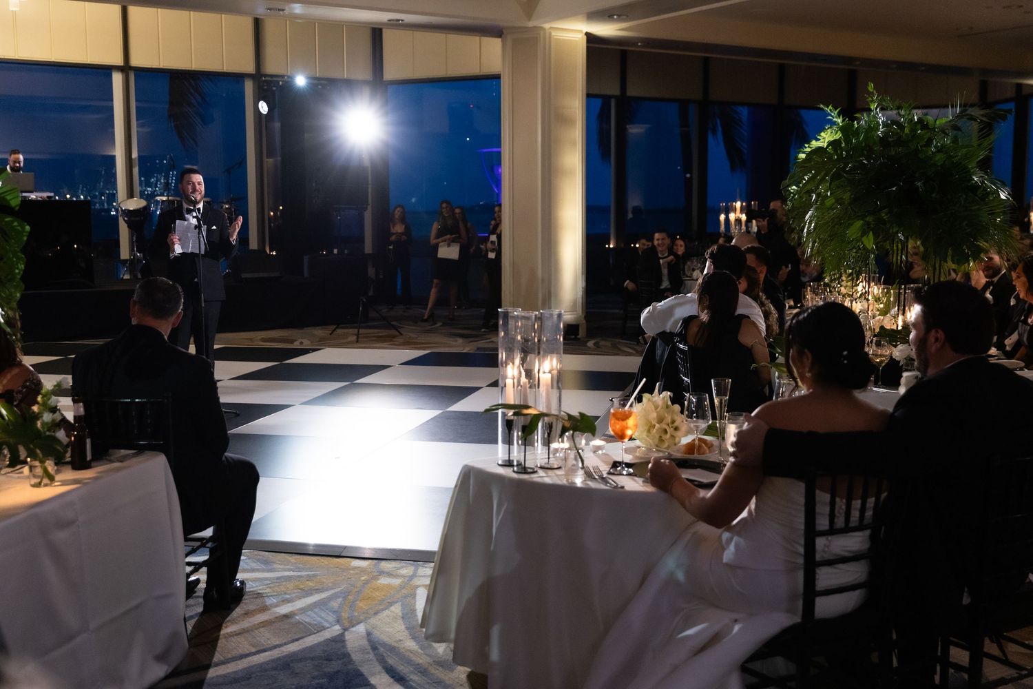 Elegant wedding reception setup with white tablecloths and ambient lighting in a room with large windows at night.