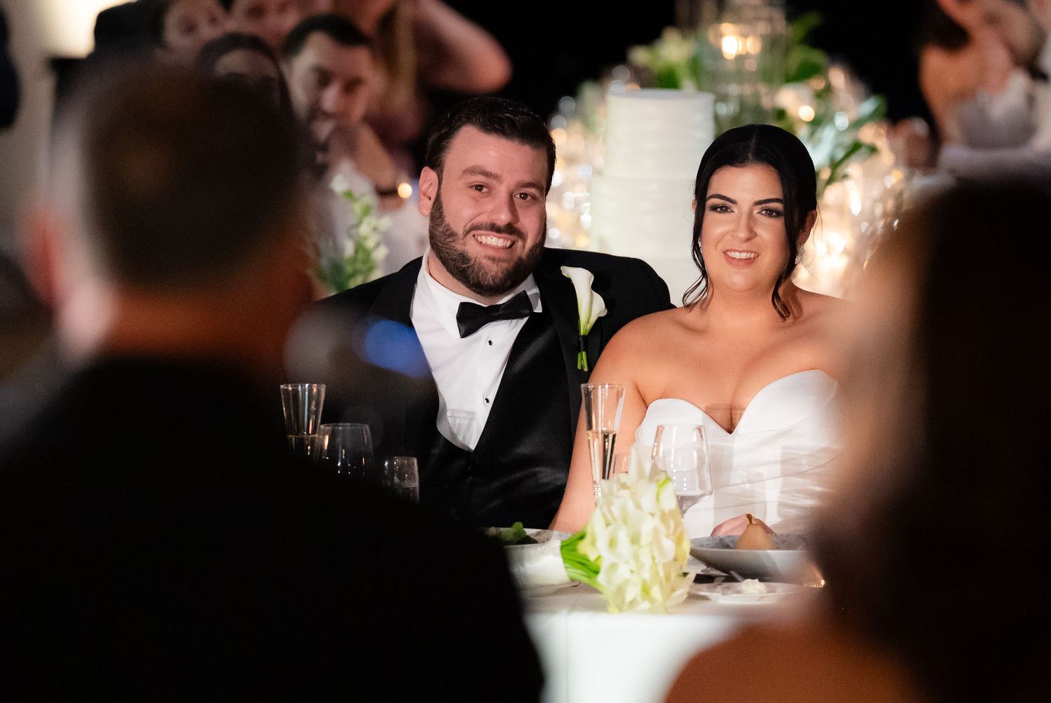 A group of wedding reception guests sit at tables lit by warm candlelight and ambient lighting during an evening celebration.