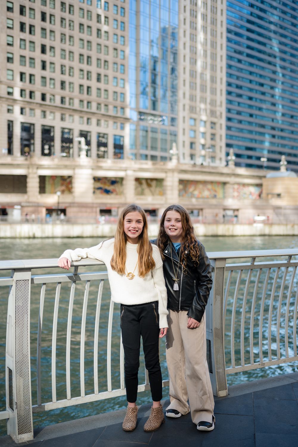 Two sisters laugh together while sitting on a railing along the Chicago River with skyscrapers in the background.