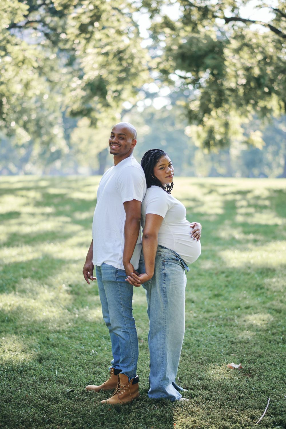 A couple stands together on a grassy hill in casual denim and white outfits on a sunny day.