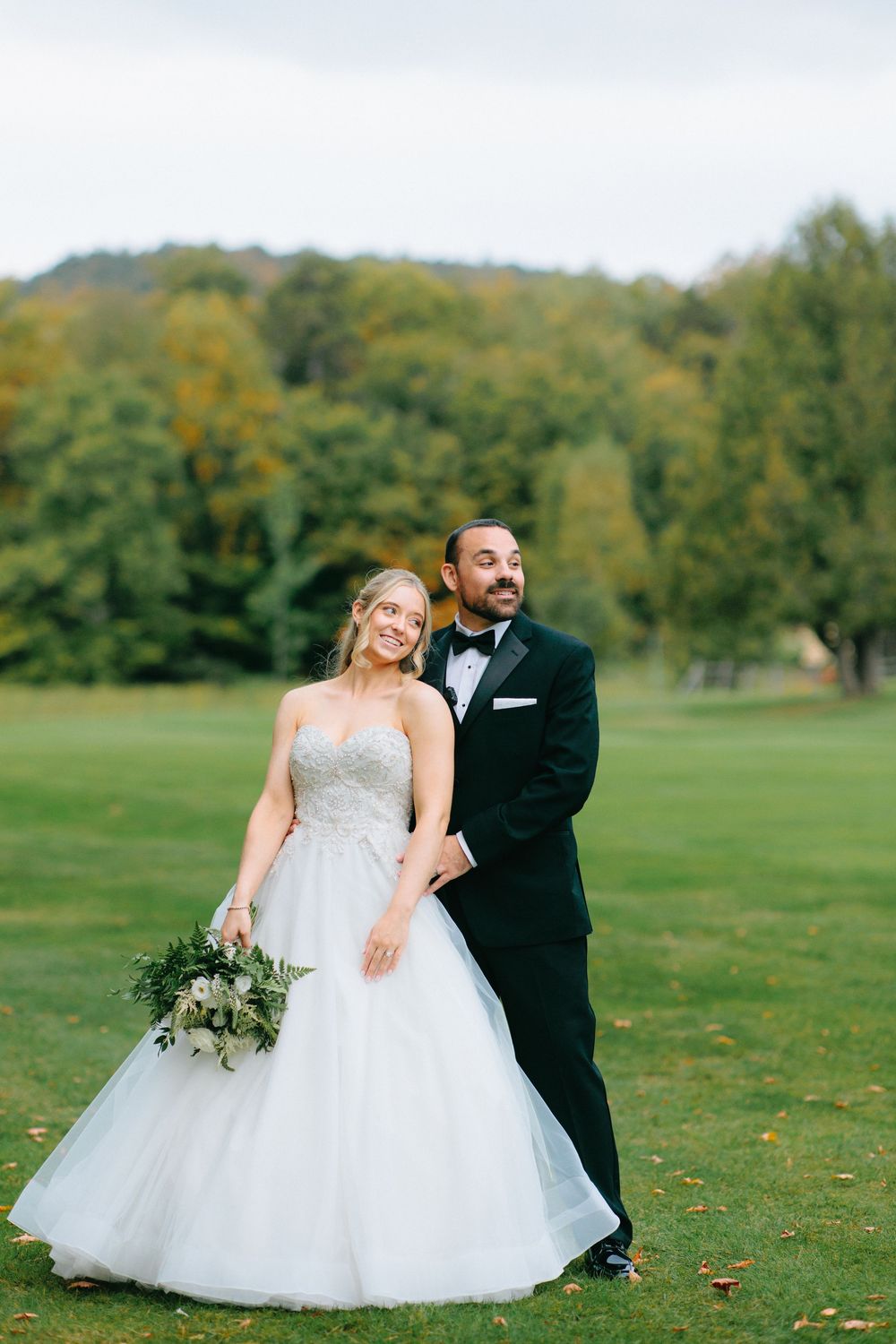A romantic wedding couple poses together on a grassy field during an autumn day.