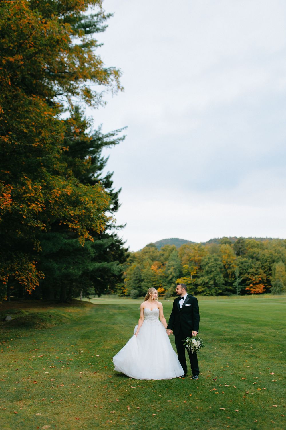 Newlyweds walk together across a scenic hilltop meadow during autumn with colorful trees in the background.