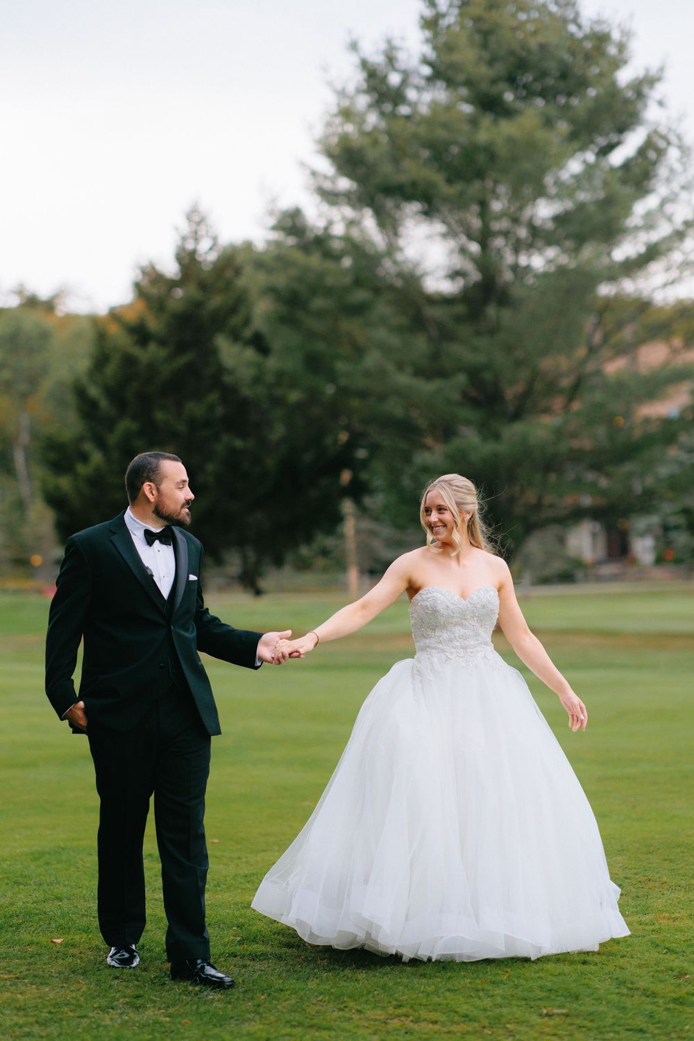Newlyweds dance playfully on a lush green lawn with pine trees in the background.