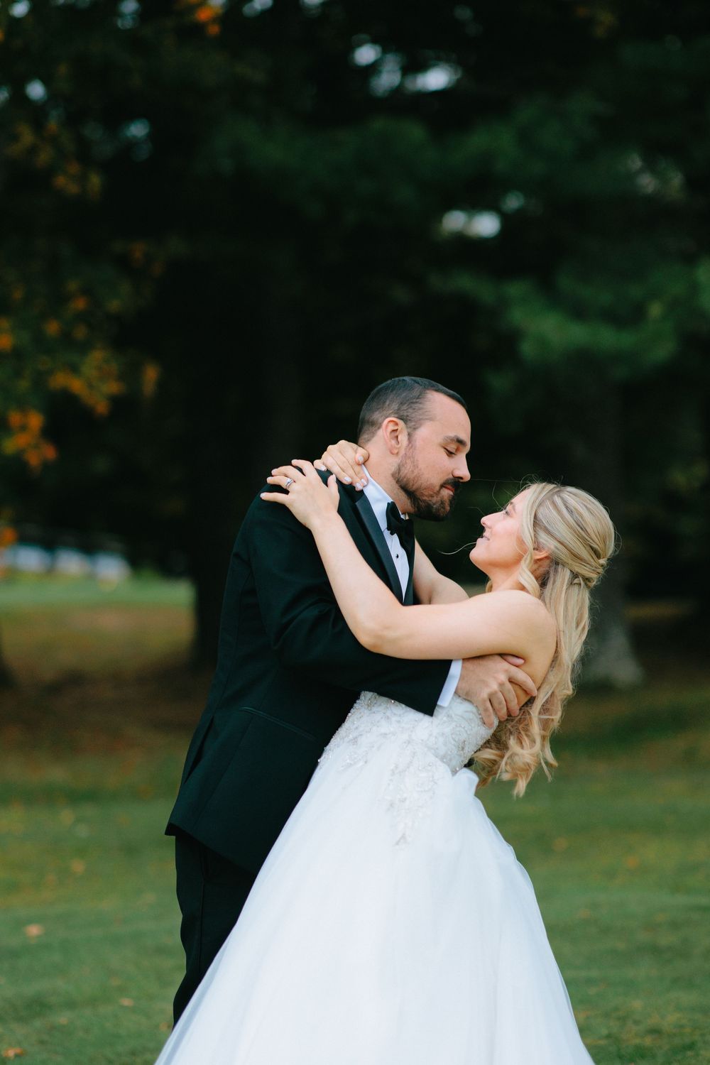 A couple shares an intimate dance pose on a scenic lawn with trees in the background.