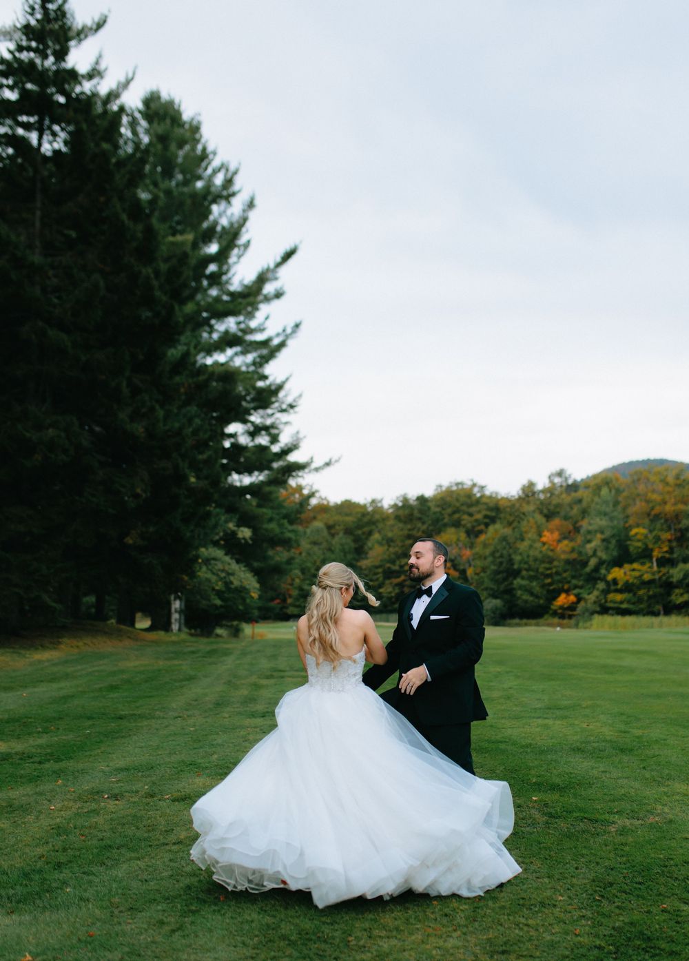 A newlywed couple twirls gracefully on a green lawn with evergreen trees in the background.