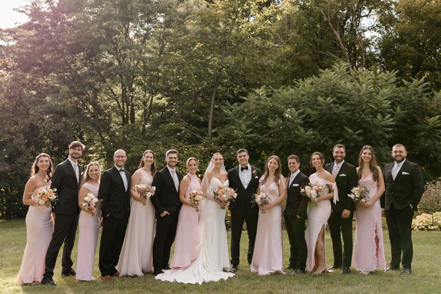 Wedding party in formal attire poses together on a lawn with trees and greenery in the background.
